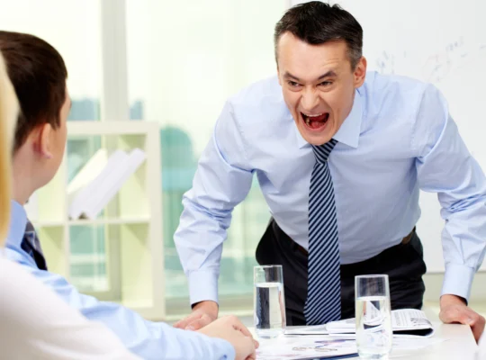 Un homme en chemise et cravate crie avec colère lors d'une réunion de travail, devant trois collègues qui écoutent attentivement. Des verres d'eau et des documents sont posés sur la table.