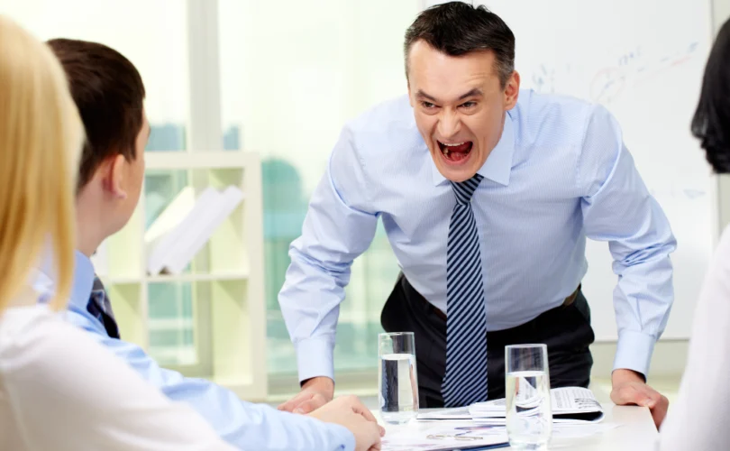 Un homme en chemise et cravate crie avec colère lors d'une réunion de travail, devant trois collègues qui écoutent attentivement. Des verres d'eau et des documents sont posés sur la table.