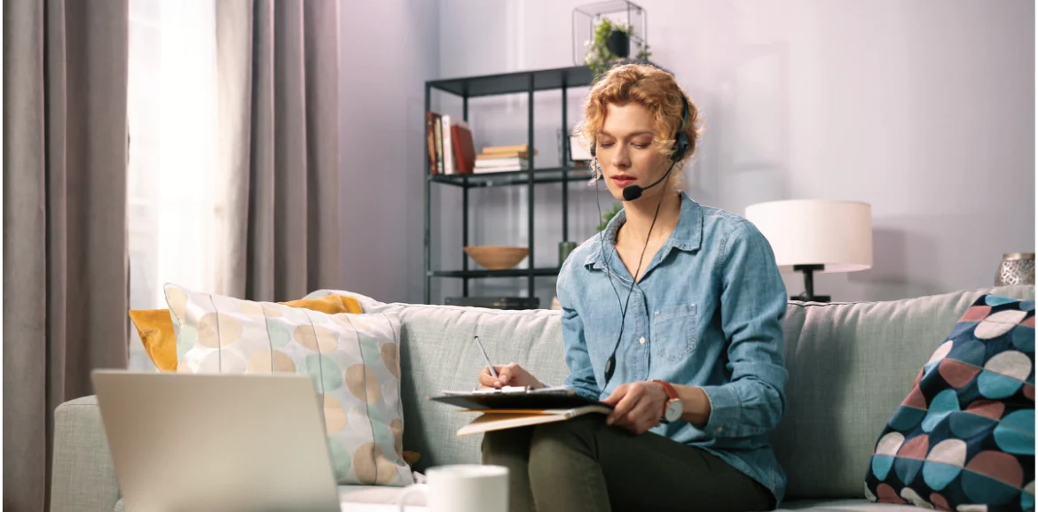 Femme avec un casque, prenant des notes lors d'une séance en ligne dans un salon confortable.
